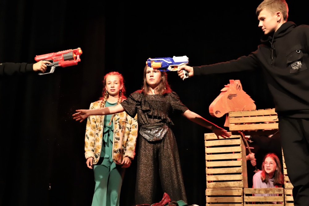 Spektakl teatralny w Ośrodku Kultury na Sali widowiskowej, 3 dziewczyny, 1 chłopak celuje plastikowym pistoletem, po prawej dziewczyna schowana za skrzynkami